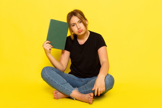A front view young female sitting in black shirt and blue jeans holding green copybook thinking depressed on yellow