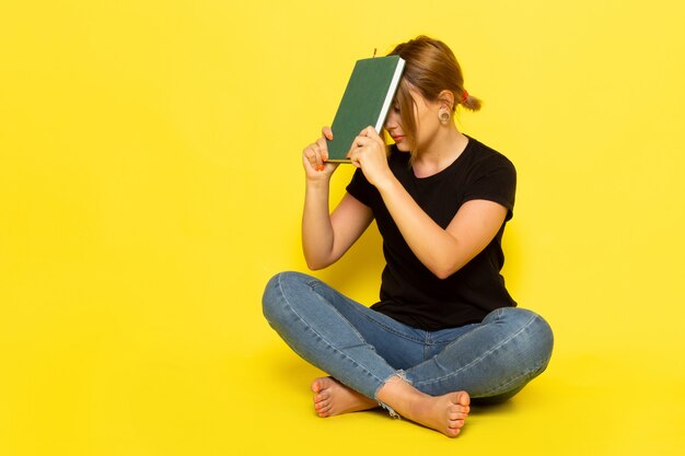 A front view young female sitting in black shirt and blue jeans holding green copybook on yellow