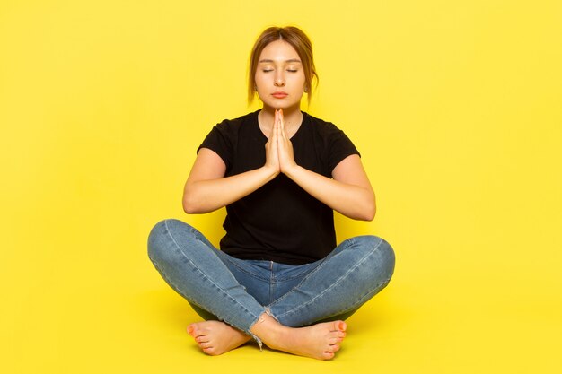 Free photo a front view young female sitting in black shirt and blue jeans meditating on yellow