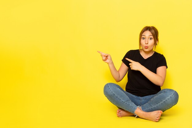 A front view young female sitting in black shirt and blue jeans posing pointing out on yellow