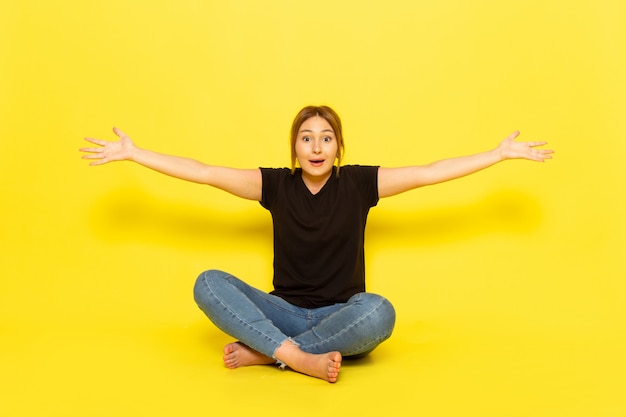 A front view young female sitting in black shirt and blue jeans spreading her arms on yellow