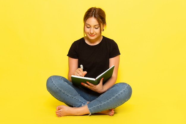 Foto gratuita una giovane donna di vista frontale che si siede in camicia nera e blue jeans che annota le note con il sorriso sul suo fronte sul colore giallo