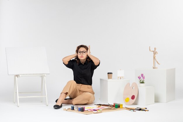 Front view young female sitting inside room with easel and paints on a white background