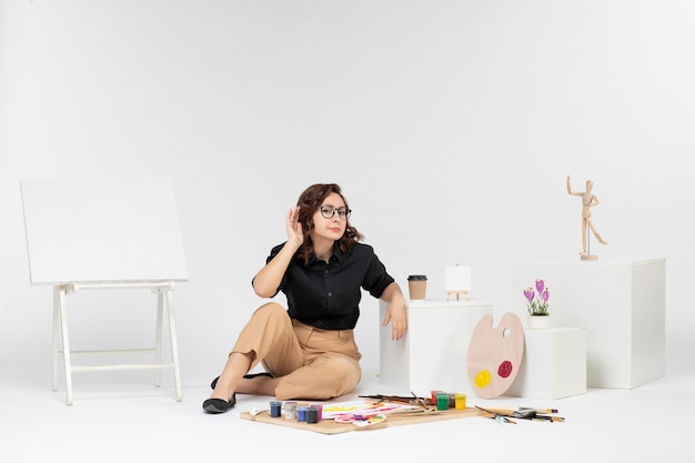 Вид спереди молодая женщина, сидящая внутри комнаты с мольбертом и красками на белом фоне