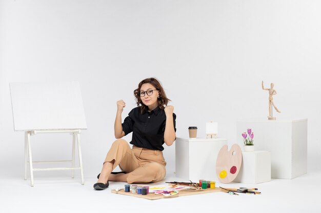 Front view young female sitting inside room with easel and paints on white background