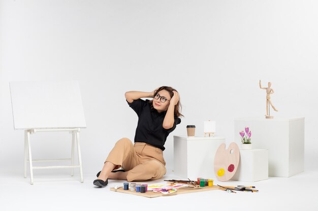 Front view young female sitting inside room with easel and paints on white background