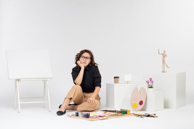 Front view young female sitting inside room with easel and paints on white background