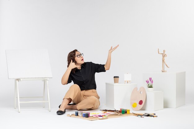 Front view young female sitting inside room with easel and paints on white background