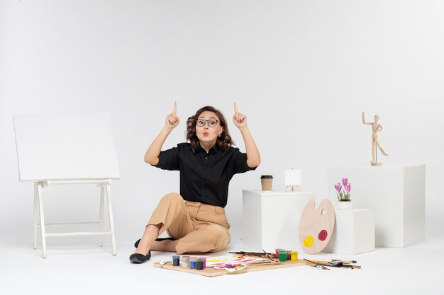 Front view young female sitting inside room with easel and paints on white background