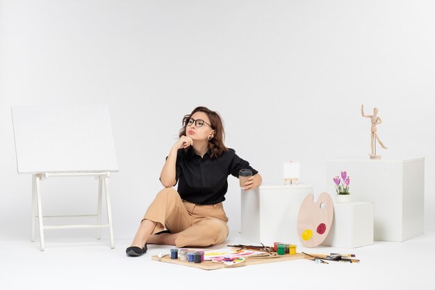 Вид спереди молодая женщина, сидящая внутри комнаты с красками и мольбертом на белом фоне