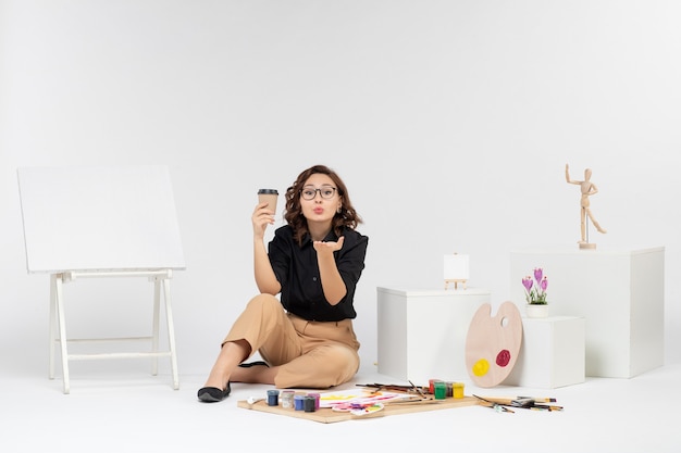 Вид спереди молодая женщина, сидящая внутри комнаты с красками и мольбертом на белом фоне