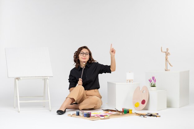 Front view young female sitting inside room with paints and easel on white background