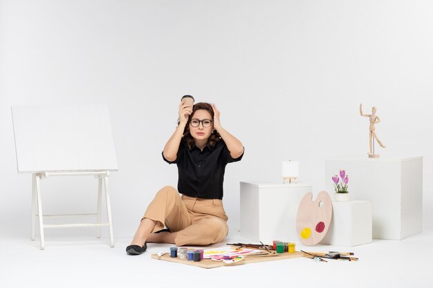 Front view young female sitting inside room with paints and easel on white desk color draw painter artist drawing art coffee