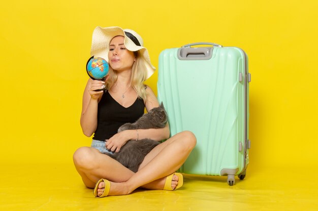 Вид спереди молодая женщина, сидящая со своей зеленой сумкой, обнимая котенка с глобусом на желтой стене, путешествие, путешествие, морское путешествие, солнце
