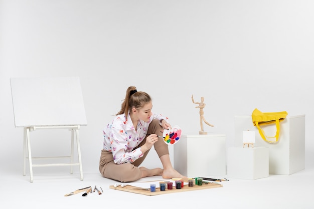 Front view young female sitting with paints and easel for drawing on a white background