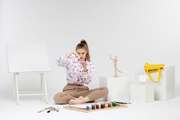 Front view young female sitting with paints and easel for drawing on the white background