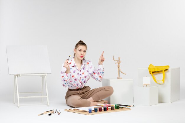 Front view young female sitting with paints and easel for drawing on white background