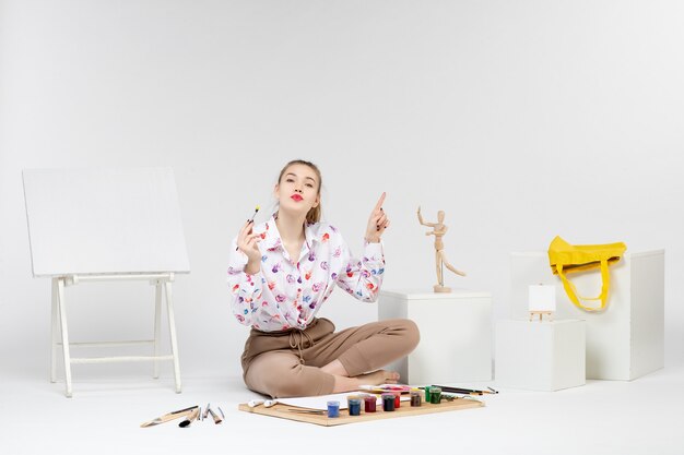 Front view young female sitting with paints and easel for drawing on white background