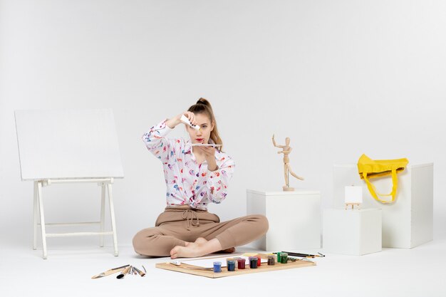 Front view young female sitting with paints and easel for drawing on white background