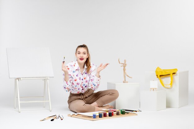 Вид спереди молодая женщина, сидящая с красками и мольбертом для рисования на белом фоне