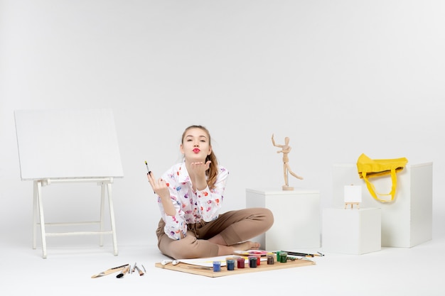 Front view young female sitting with paints and easel for drawing on white background