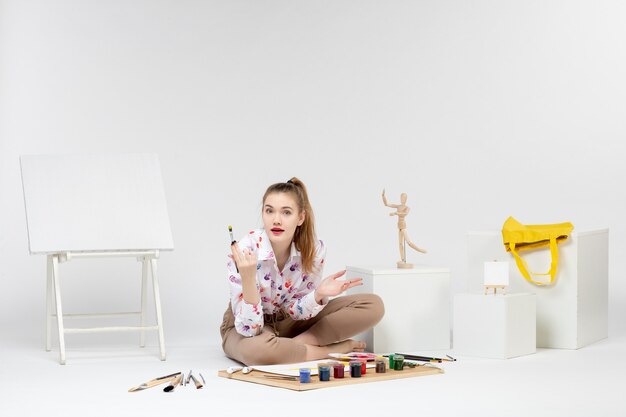 Front view young female sitting with paints and easel for drawing on white background