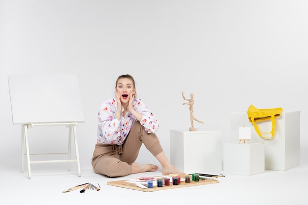 Front view young female sitting with paints easel and paint brushes excited on white background