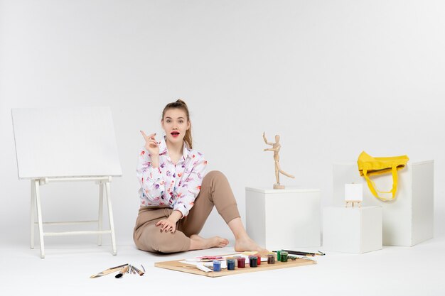 Front view young female sitting with paints easel and paint brushes on a white background