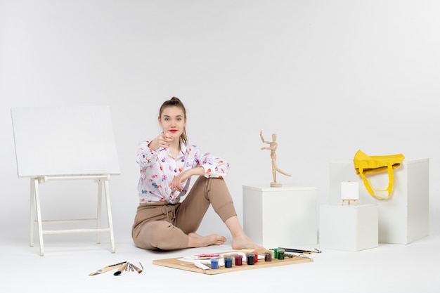 Front view young female sitting with paints easel and paint brushes on the white background