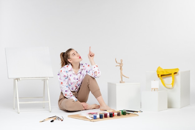 Front view young female sitting with paints easel and paint brushes on white background