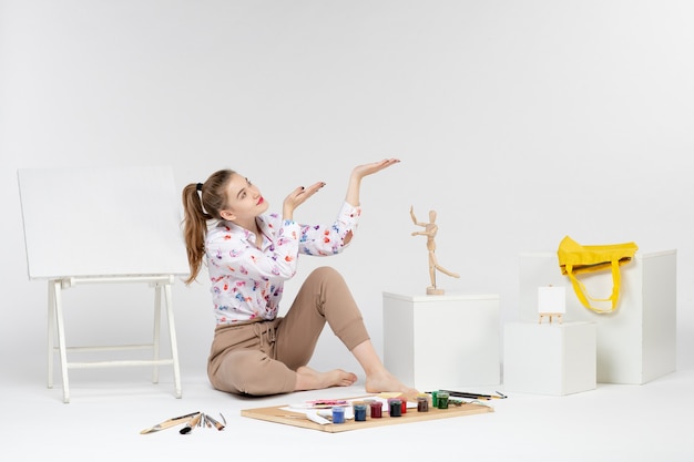 Front view young female sitting with paints easel and paint brushes on white background