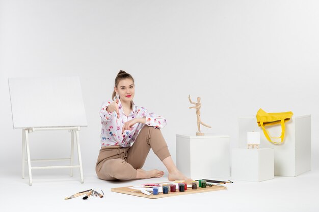 Front view young female sitting with paints easel and paint brushes on white background