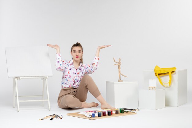 Front view young female sitting with paints easel and paint brushes on white background