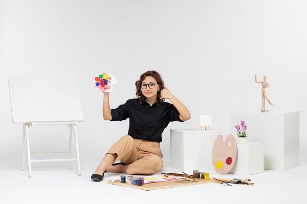 Front view young female sitting with paints and easel on white background