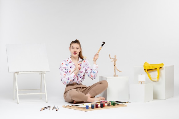 Front view young female sitting with paints holding paint brushes on the white background