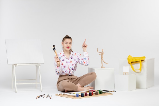 Front view young female sitting with paints holding paint brushes on the white background