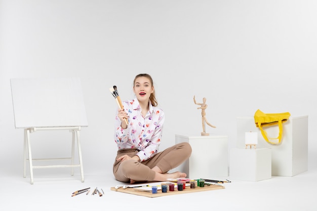 Front view young female sitting with paints holding paint brushes on white background