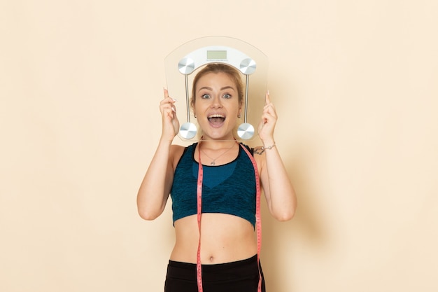 Front view young female in sport outfit holding scales on a light-white wall fit body sport beauty health exercises