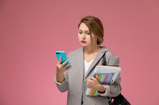 ピンクの背景のレッスン大学大学の研究で電話を使用して本を持ってポーズをとる灰色のコートの若い女子学生