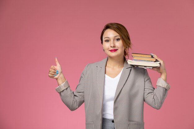 Foto gratuita vista frontale giovane studentessa in cappotto grigio in posa tenendo libri con il sorriso sullo sfondo rosa lezioni di studio universitario