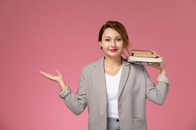 Free photo front view young female student in grey coat posing and holding copybooks with smile on pink background lessons university college study