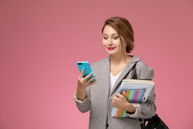 Front view young female student in grey coat posing and using phone on the pink background lessons university college study