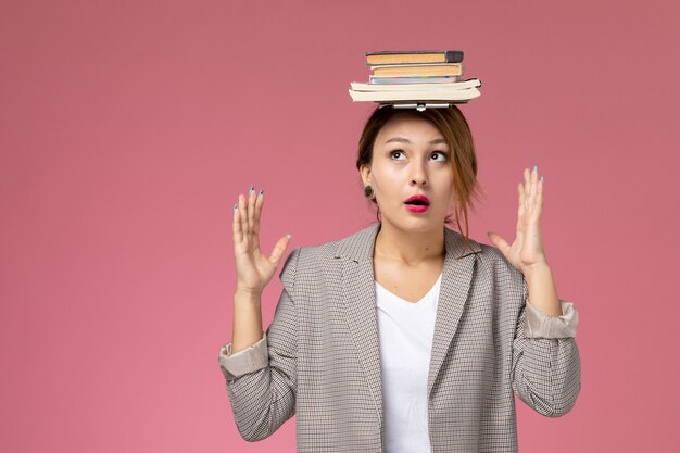 분홍색 배경 수업 대학 대학 연구 책에 그녀의 머리에 책과 함께 포즈 회색 코트에 전면보기 젊은 여성 학생