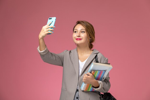 Front view young female student in grey coat posing with copybooks and taking a selfie on pink background lessons university college study