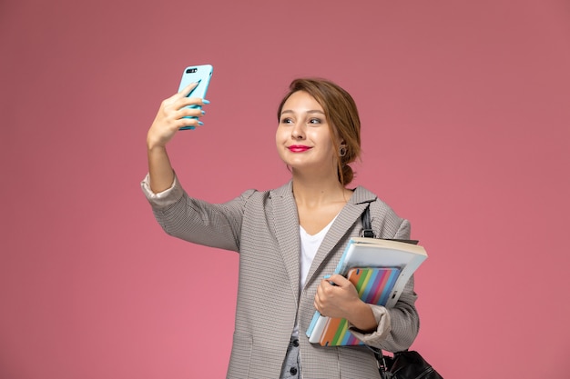 Front view young female student in grey coat posing with copybooks and taking a selfie on pink background lessons university college study