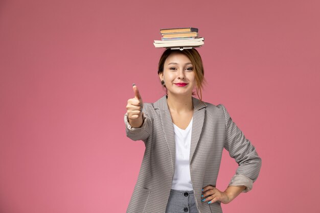 Front view young female student in grey coat with copybooks on her head smiling on pink background lessons university college study