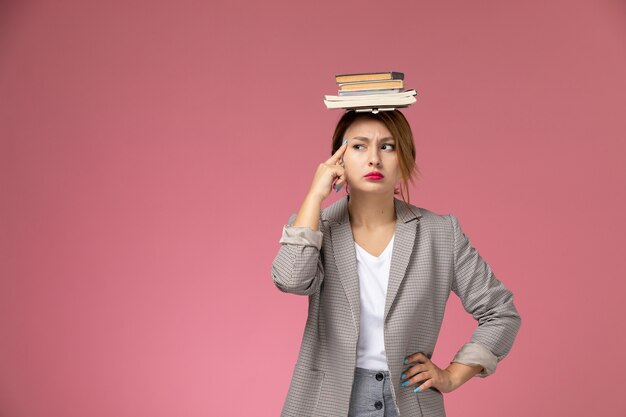 Front view young female student in grey coat with copybooks on her head with thinking expression on the pink background lessons university college study