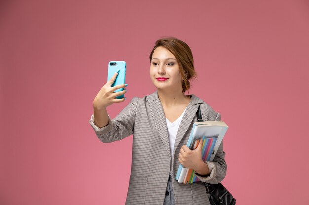 Front view young female student in grey coat with copybooks taking a selfie on pink background lessons university college study