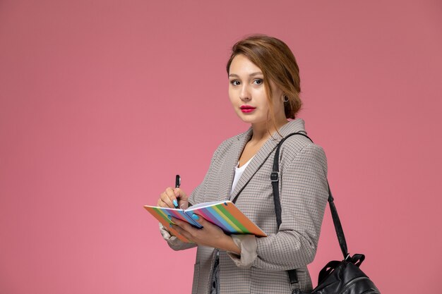 Front view young female student in grey coat with copybooks writing down on pink background lessons university college study