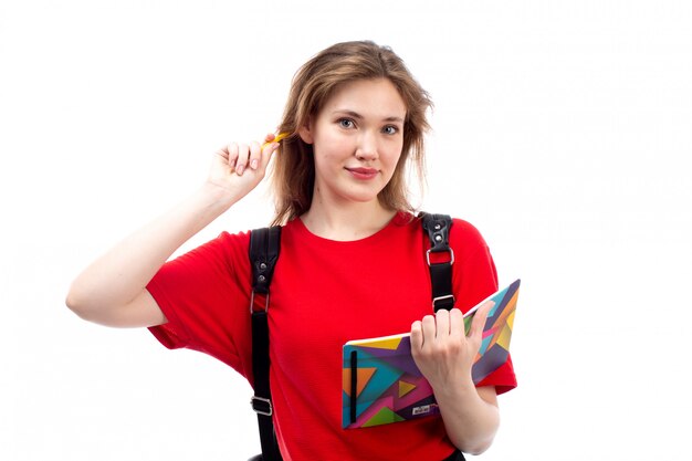 Вид спереди молодая студентка в красной рубашке черная сумка с копирайтом файлы улыбаясь, записывая на белом